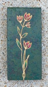 Clay paver with stylized gold-outlined flowers on a green background engraved and painted on it.