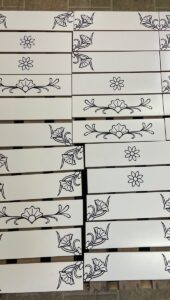 Assembly of 4in. x 16in. white subway tiles engraved and painted black prepared for installation Group of white subway tiles engraved with various designs prepared for installation.